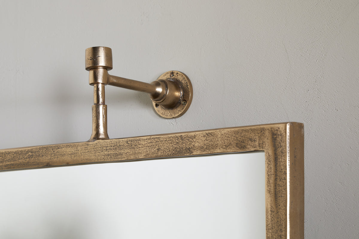 Close-up of the nkuku Visala Swivel Wall Mirror in Antique Brass, featuring a rectangular gold metallic frame and mounted on a light gray wall with a cylindrical brass bracket at the upper left corner.
