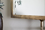 A close-up of the nkuku Visala Swivel Wall Mirror in antique brass shows its textured gold frame reflecting green foliage. A brown ribbed vase with leaves is at the left edge, all set against a light-colored wall.