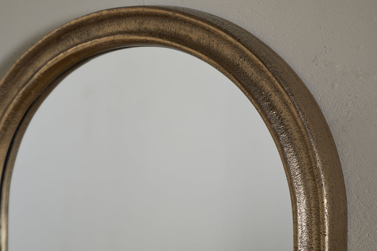 A close-up of the upper left portion of the Mbwa Oval Mirror - Antique Brass by nkuku shows its textured, metallic frame. The decorative mirror reflects a plain light gray wall, highlighting the frame’s curved edge and rough finish.