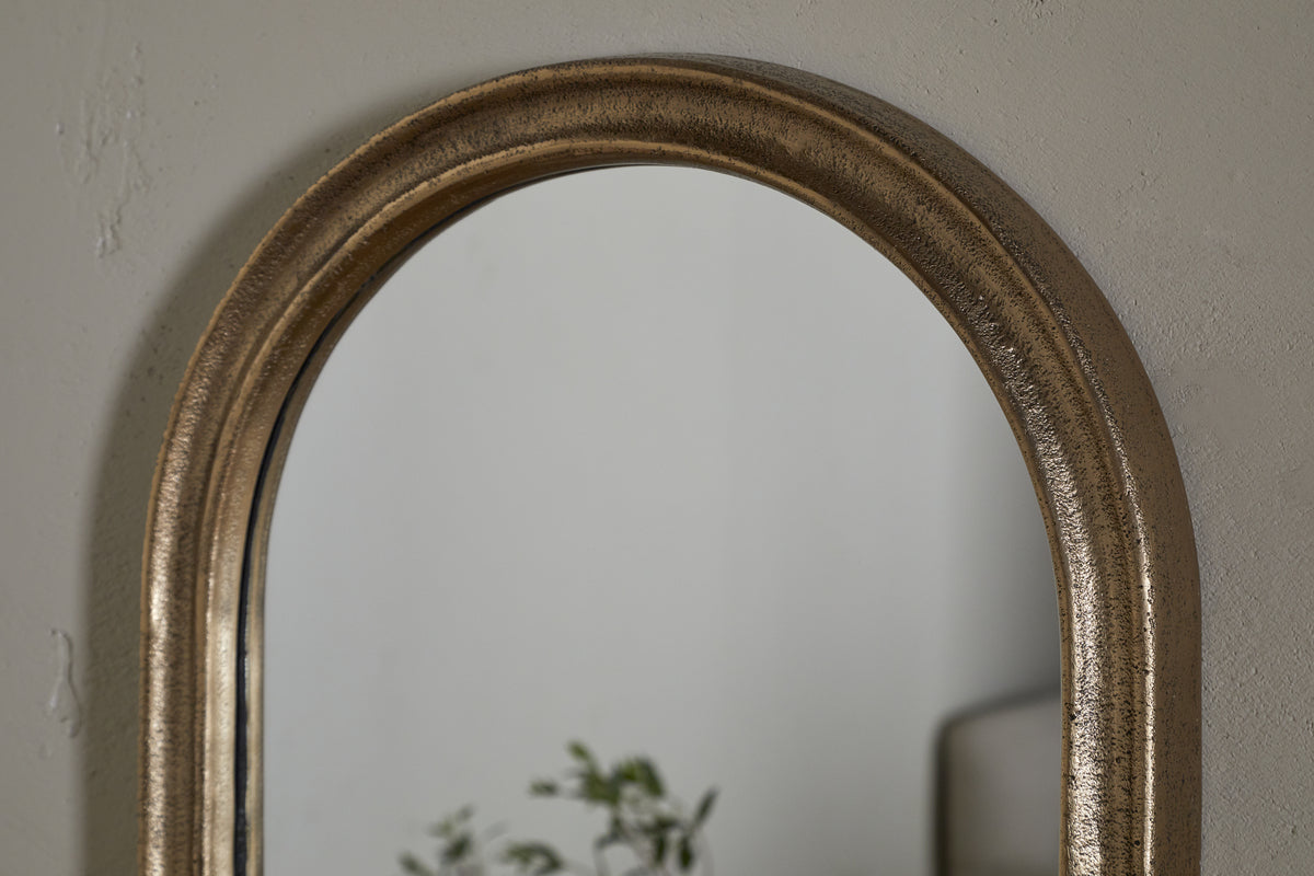 A close-up of the nkuku Mbwa Oval Mirror in Antique Brass, featuring a textured frame on a light gray wall. The mirror reflects blurred green plant leaves and part of neutral furniture.
