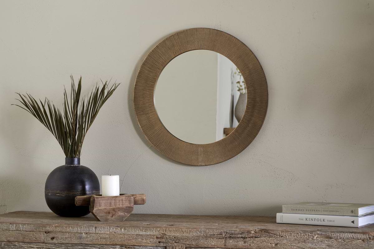 The Putali Etched Mirror - Antique Brass by nkuku hangs on a beige wall above a rustic wood console table topped with a black vase of dried grass, a white candle on wood, and stacked books for a minimal, stylish scene.