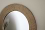 Close-up of the Putali Etched Mirror - Antique Brass by nkuku, showcasing its textured gold-toned frame with fine vertical grooves. The round mirror reflects thin green plant leaves against a plain off-white wall.
