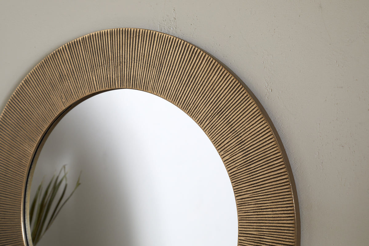 Close-up of the Putali Etched Mirror - Antique Brass by nkuku, showcasing its textured gold-toned frame with fine vertical grooves. The round mirror reflects thin green plant leaves against a plain off-white wall.