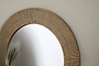 Close-up of the Putali Etched Mirror - Antique Brass by nkuku, showcasing its textured gold-toned frame with fine vertical grooves. The round mirror reflects thin green plant leaves against a plain off-white wall.