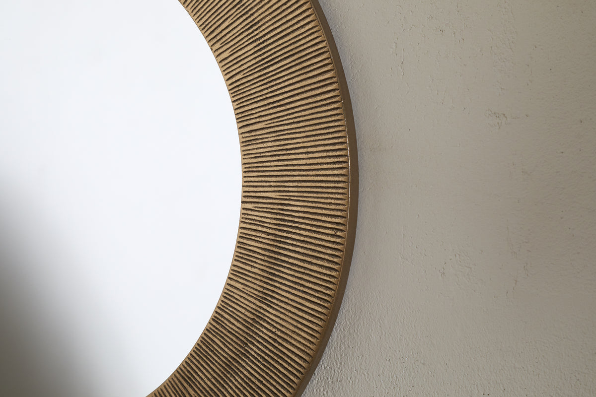 Close-up of the Putali Etched Mirror - Antique Brass by nkuku, featuring a sculptural, wide wooden frame with vertical grooves. The round mirror hangs on a light wall, reflecting a clean white space.