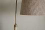 Close-up of the nkuku Kera Wood & Metal Floor Lamp in Antique Brass & Natural, showing part of its brass arm and textured beige shade against a light-colored, cracked wall.