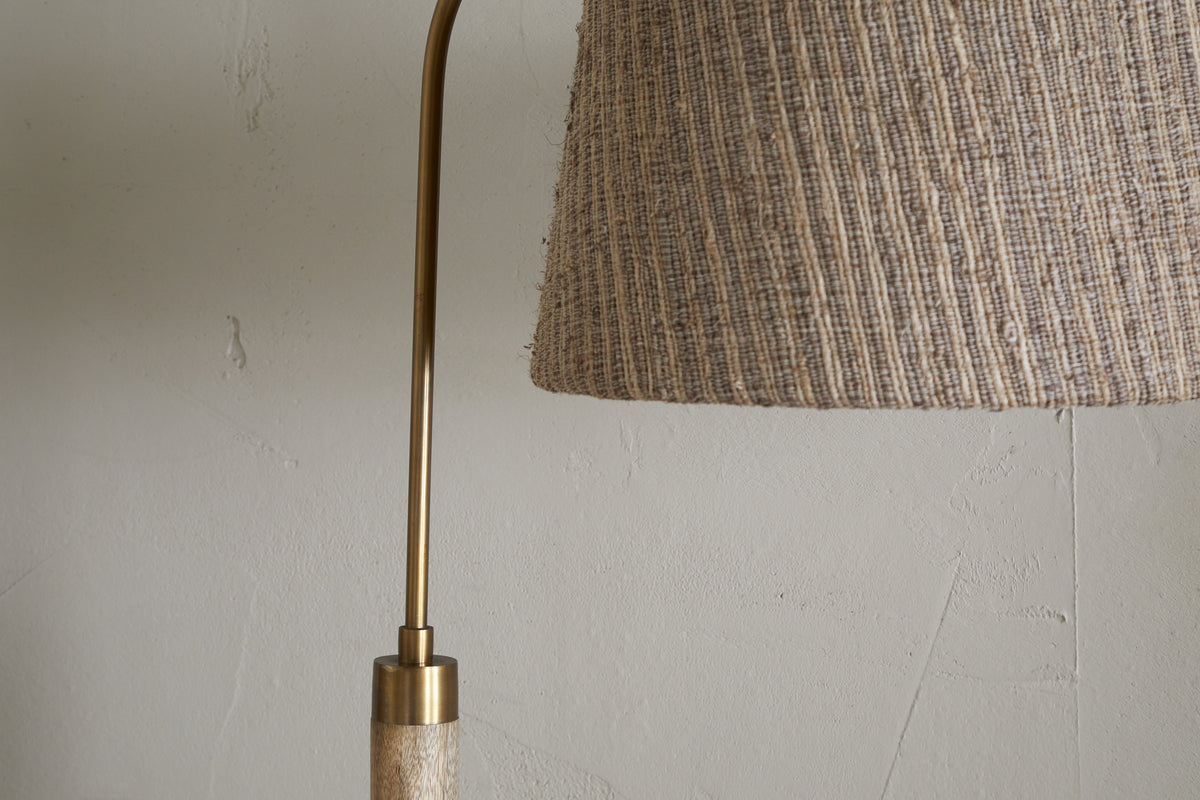 Close-up of the nkuku Kera Wood & Metal Floor Lamp in Antique Brass & Natural, showing part of its brass arm and textured beige shade against a light-colored, cracked wall.