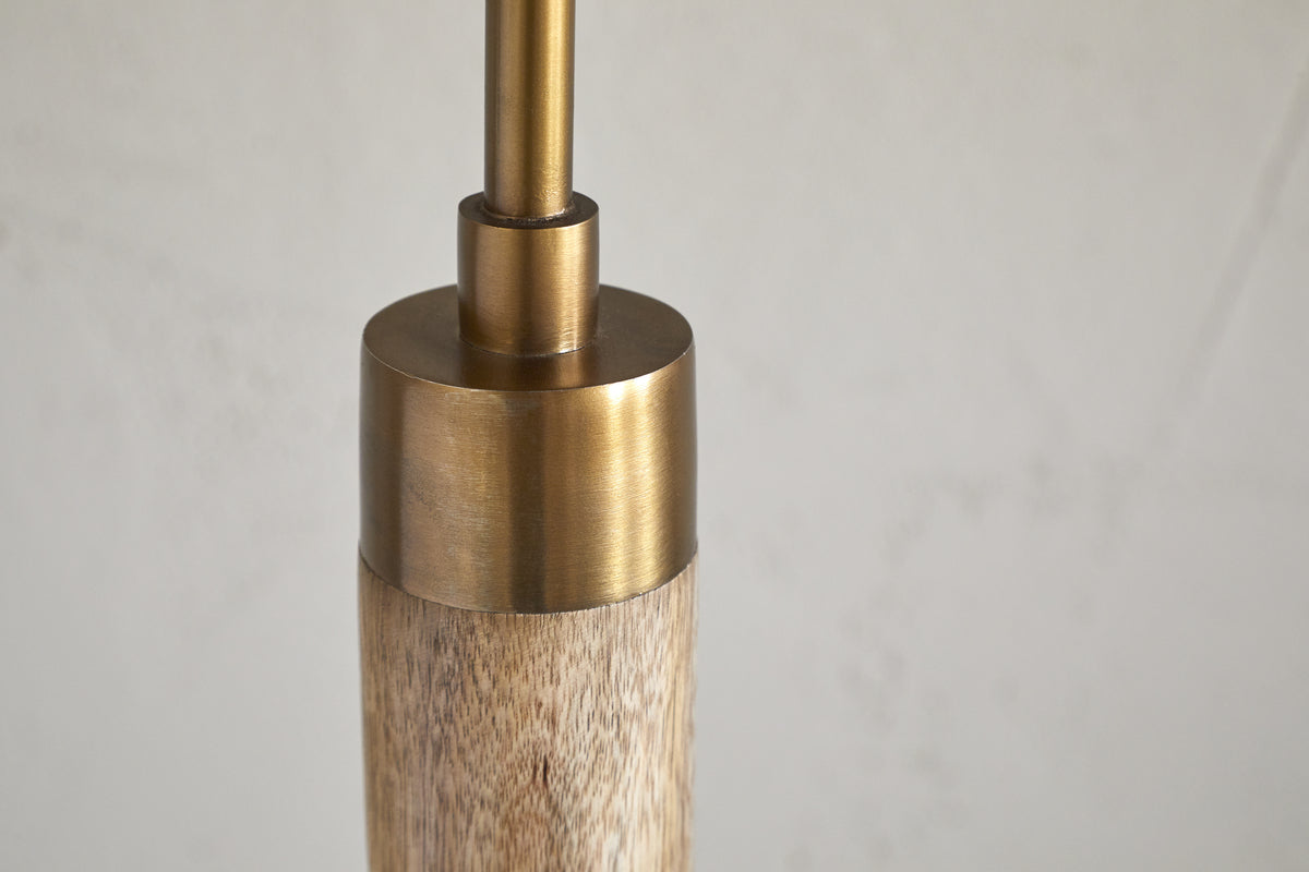 Close-up of the nkuku Kera Wood & Metal Floor Lamp - Antique Brass & Natural highlights its cylindrical mango wood base and slender brushed brass rod, set against a subtly textured, pale background.