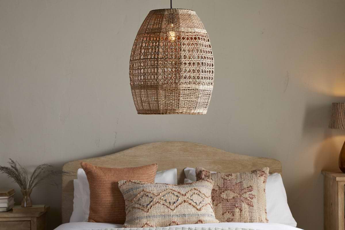 The Shawa Rattan Lampshade by nkuku hangs above a wooden bed with a curved headboard, dressed in white bedding and earthy-toned, patterned throw pillows. A matching handwoven shade and decorative vase accent the nightstand.