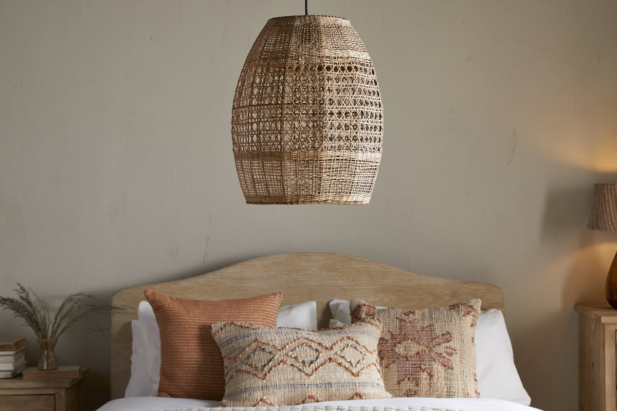 The Shawa Rattan Lampshade by nkuku hangs above a bed with a light wood headboard. Textured earth-toned pillows add warmth, while a wooden nightstand with a lamp and vase sits to the right.