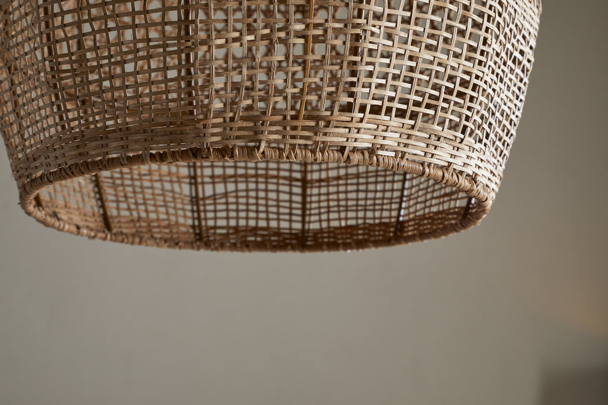 Close-up of the nkuku Shawa Rattan Lampshade - Natural, featuring intricate handwoven crisscross patterns and a natural beige finish. The blurred background enhances the texture and craftsmanship of this elegant lighting accent.