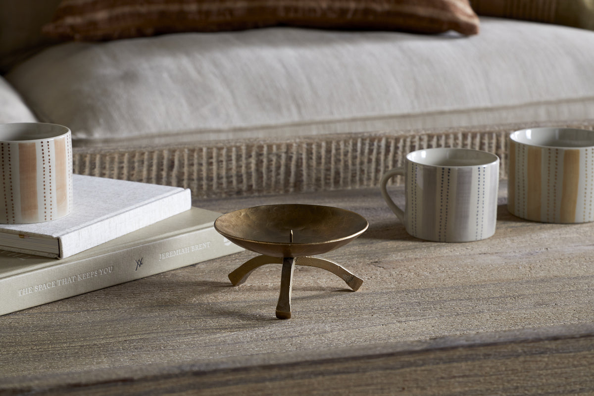 The nkuku Kalik Candle Holder in antique brass, featuring three legs, is displayed on a wooden table beside two patterned ceramic mugs and two stacked light-covered books. A beige cushioned sofa with a brown pillow adds warmth in the background.