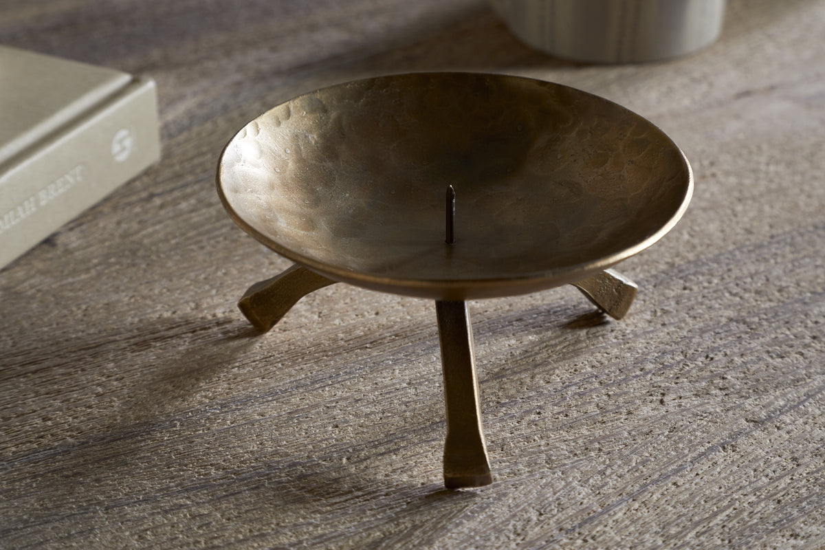 The Kalik Candle Holder by nkuku, crafted in antique brass with three angled legs and a central spike for securing a pillar candle, stands on a textured wooden surface near a beige book and a striped cylindrical object.