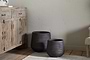 Two Lmaga Metal Grooved Planters by nkuku in aged black, one large and one small, sit on a wooden floor near a rustic cabinet, creating a cozy minimalist space perfect for stylish indoor plants.