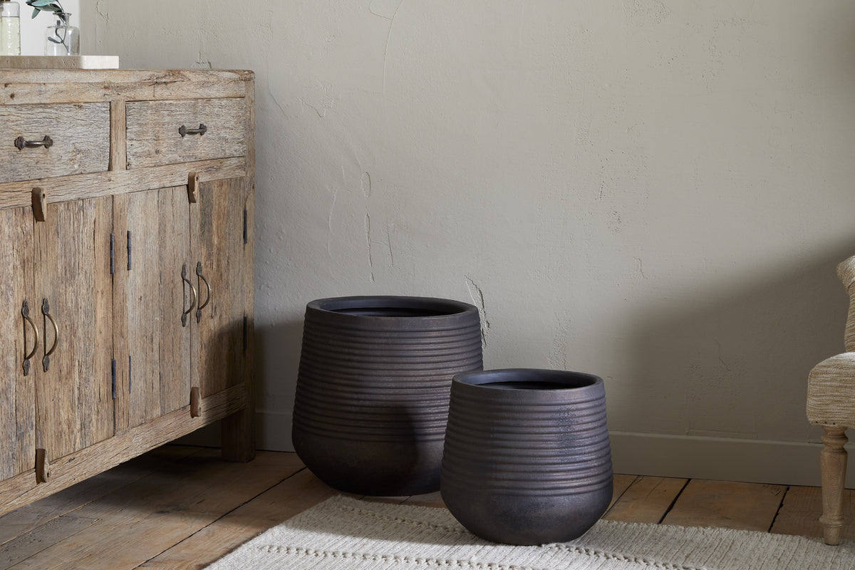 Two Lmaga Metal Grooved Planters by nkuku in aged black, one large and one small, sit on a wooden floor near a rustic cabinet, creating a cozy minimalist space perfect for stylish indoor plants.