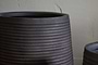Close-up of the nkuku Lmaga Metal Grooved Planter in Aged Black, featuring textured horizontal lines. The large pot stands before a rough beige wall, with part of a smaller matching planter visible to the right.