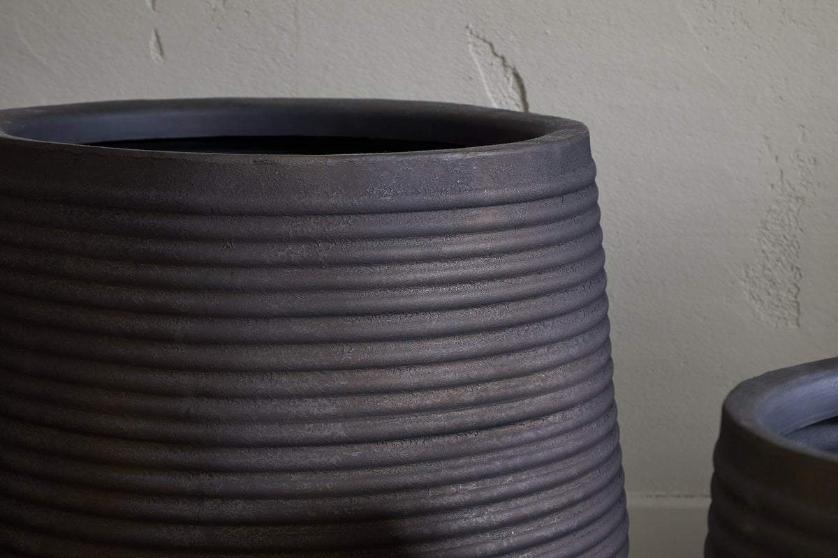 Close-up of the nkuku Lmaga Metal Grooved Planter in Aged Black, featuring textured horizontal lines. The large pot stands before a rough beige wall, with part of a smaller matching planter visible to the right.