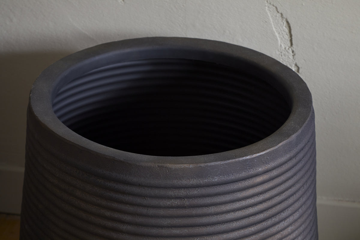 The nkuku Lmaga Metal Grooved Planter in aged black features thick walls, a smooth rounded rim, and a matte finish. Its large, empty form stands out near a light-colored, slightly textured wall—ideal for displaying houseplants.