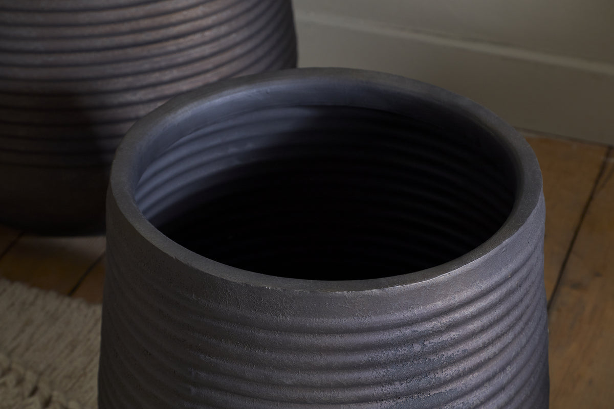 Close-up of the nkuku Lmaga Metal Grooved Planter in Aged Black, its stylish horizontal ridges visible as it sits on a wooden floor beside a textured beige rug. Another similar planter appears blurred in the softly lit background.