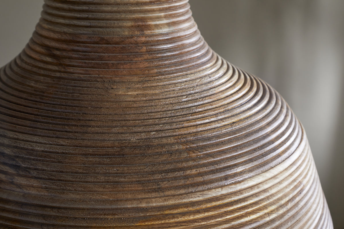 Close-up of the Nadi Grooved Wood Pendant by nkuku, featuring rounded, tapering mango wood with concentric grooves and an antique brass finish. The polished brown grain stands out against a soft beige and gray backdrop.