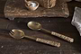 The Abani Salad Servers by nkuku—antique brass with woven rattan handles—rest on a rustic wood table, joined by a cutting board, seagrass basket, salt bowl, and cloth napkin.