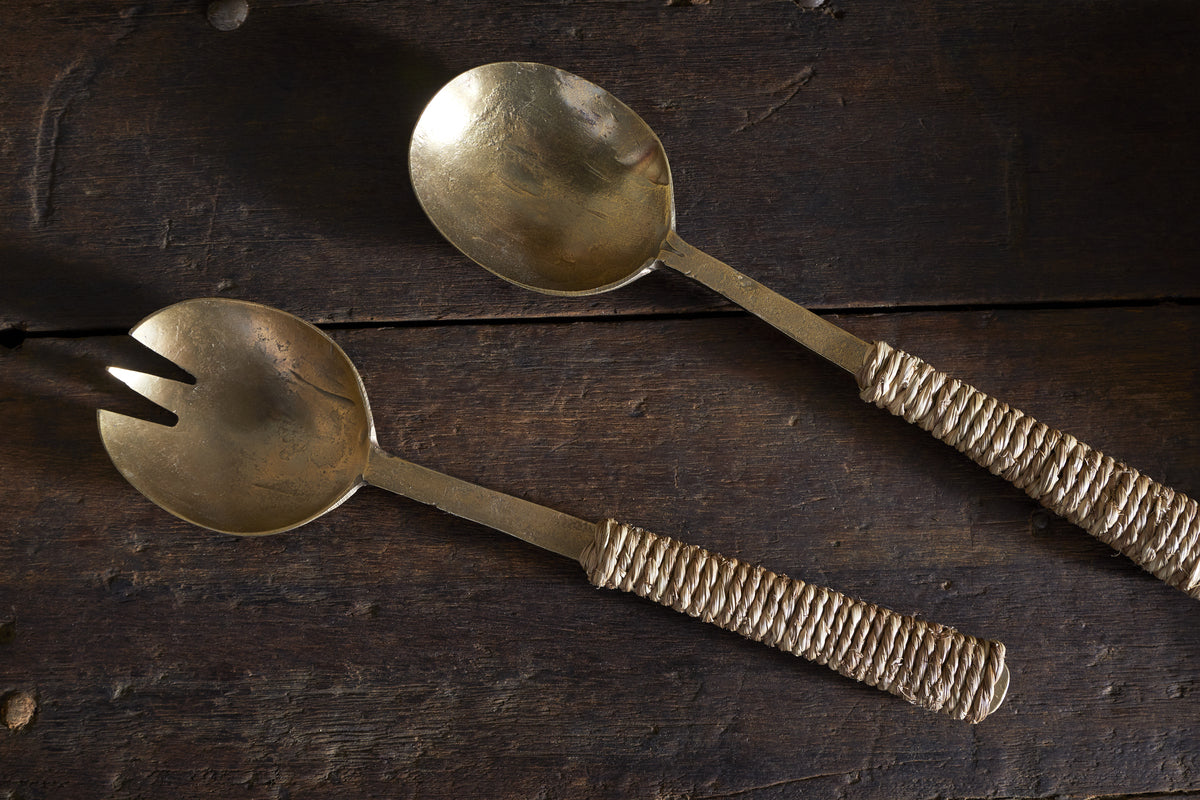 The Abani Salad Servers by nkuku—an antique brass spoon and spork—rest on a dark wooden surface, their seagrass-wrapped textured handles lending this handcrafted tableware a rustic, artisanal touch.