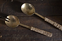 The Abani Salad Servers by nkuku—an antique brass spoon and spork—rest on a dark wooden surface, their seagrass-wrapped textured handles lending this handcrafted tableware a rustic, artisanal touch.