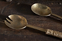 The nkuku Abani Salad Servers in antique brass feature a spoon and two-tined fork-spoon with square handles. Handcrafted with seagrass twine on one handle, their slightly tarnished surfaces add rustic charm.
