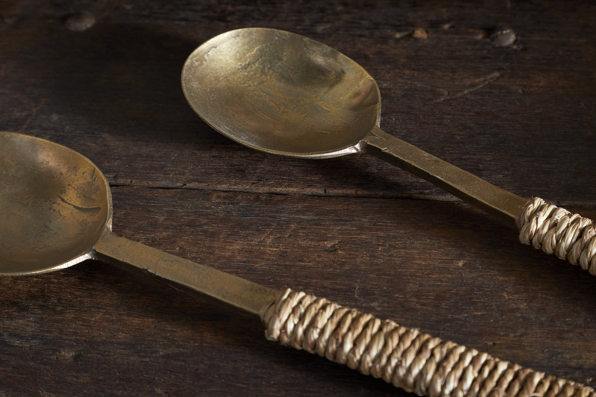 The Abani Salad Servers by nkuku feature antique brass and square handles wrapped in natural twine, their matte finish showcasing handcrafted charm as they rest on rustic, weathered wood.