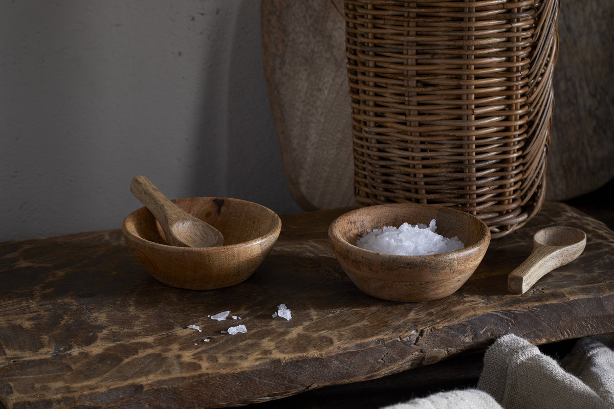 The nkuku Daruka Salt & Pepper Dipping Bowls with Spoons - Natural rest on a rustic wooden surface, one cradling coarse sea salt, the other empty but for a spoon. Scattered salt crystals and cozy table accents enhance the inviting scene.
