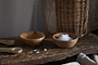The nkuku Daruka Salt & Pepper Dipping Bowls with Spoons – Natural, paired with a wicker basket and beige cloth, add warmth and earthiness to your table as inviting wooden accessories.