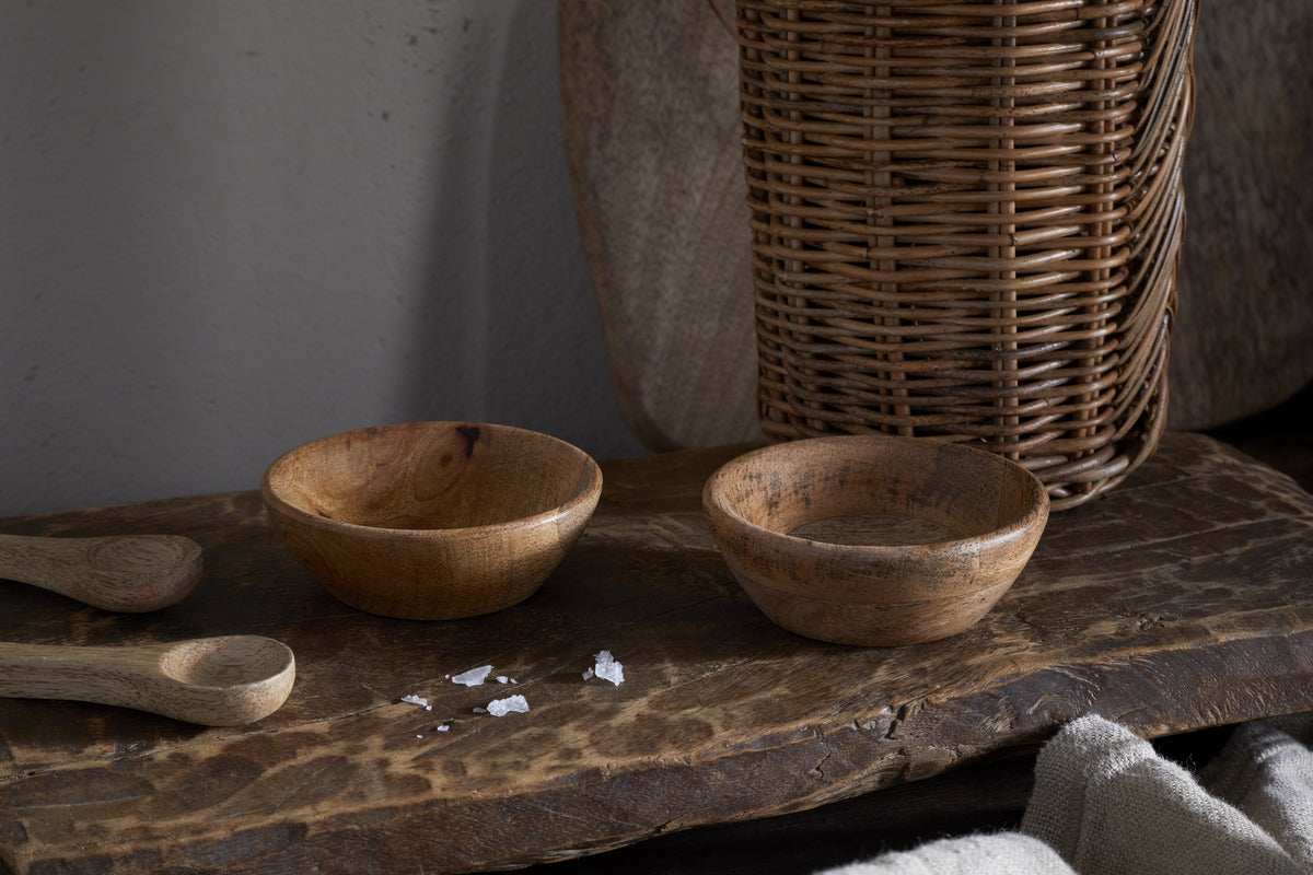 The nkuku Daruka Salt & Pepper Dipping Bowls with Spoons - Natural are shown on a rustic surface beside coarse salt, with a wicker basket and beige cloth nearby—ideal for serving condiments stylishly.