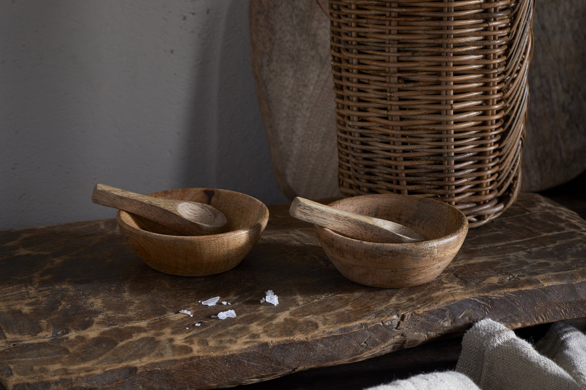 The nkuku Daruka Salt & Pepper Dipping Bowls with Spoons – Natural sit on a rustic surface. Coarse salt crystals and warm table accessories like a wicker basket and textured boards complete this inviting, natural set.