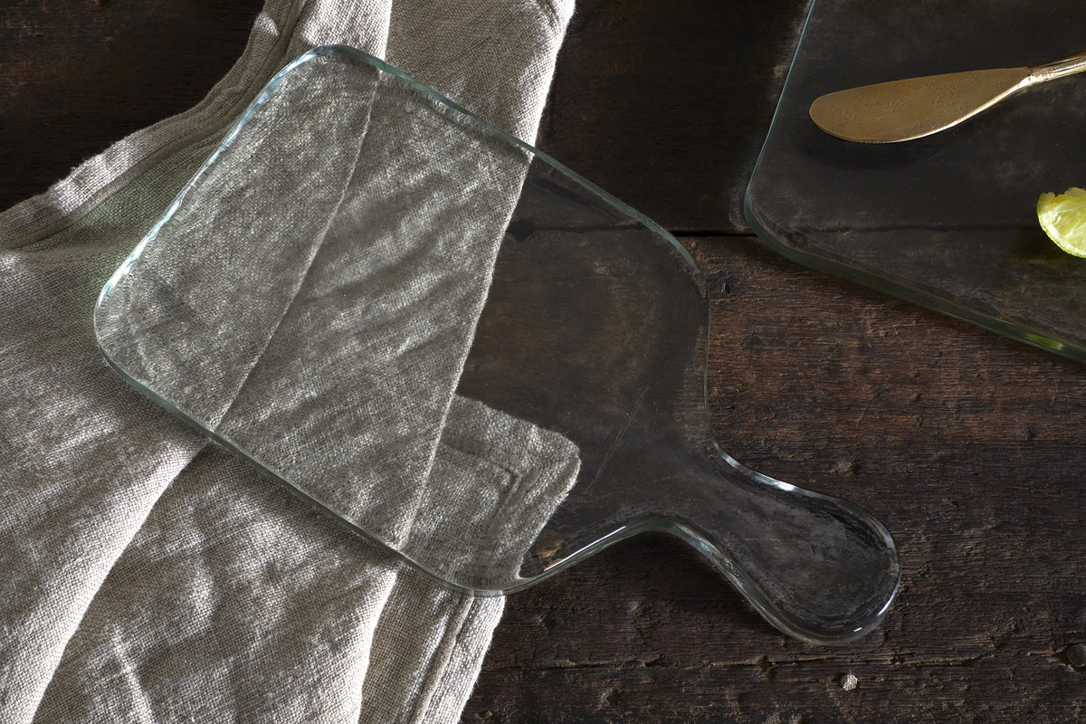 The nkuku Komal Serving Platter - Clear with a handle sits on a beige linen atop dark wood, while another stylish platter with a gold butter knife and lime slice appears in the top right corner.