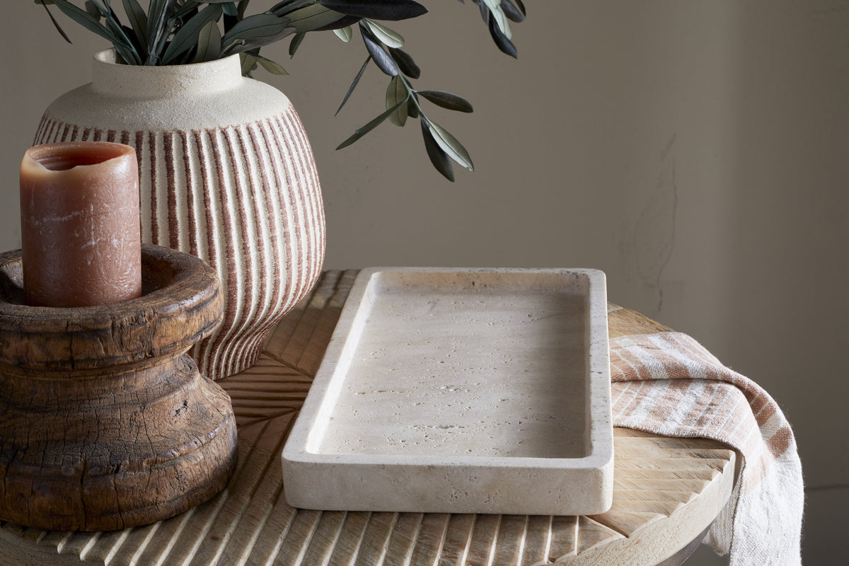 A beige ceramic vase with green leaves, a brown pillar candle on a rustic wooden stand, and the nkuku Ganda Travertine Vanity Tray in cream sit atop a round table with a beige-striped cloth, creating a softly lit, minimalistic ambiance.