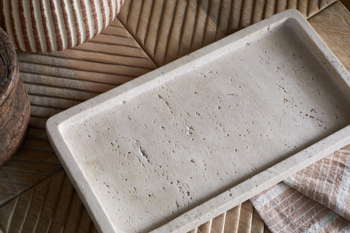 The nkuku Ganda Travertine Vanity Tray in Cream, featuring a rough, porous finish, rests on a wooden geometric surface beside a striped beige cloth, round wooden container, and woven basket for a neutral bathroom organiser look.
