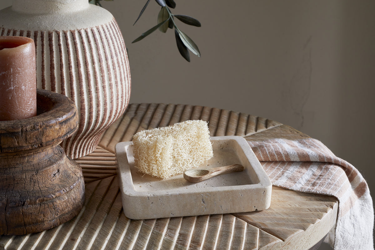 A natural loofah and a small wooden spoon sit on the cream Ganda Travertine Vanity Tray by nkuku, atop a carved wooden table. Nearby are a brown ribbed candle, pink-striped vase, and folded beige cloth in gentle light and soft shadows.