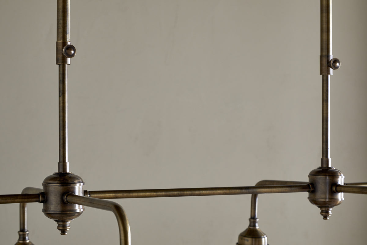 Close-up of the nkuku Kakira Chandelier Pendant in Antique Brass & Clear, featuring visible rods and joints, clean lines, metal hardware, and a muted finish—all set against a plain beige wall for an industrial yet elegant look.