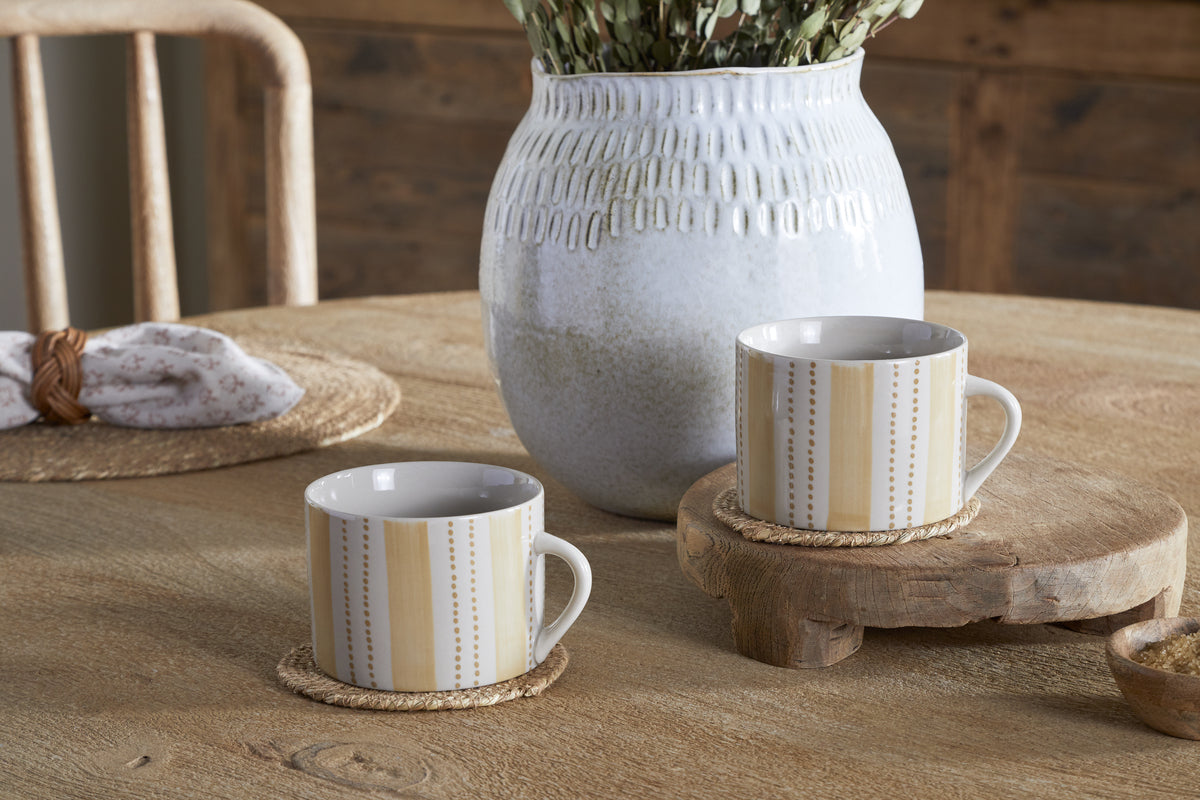 Two Patthar Mugs in Cream & Sand by nkuku rest on round coasters atop a wooden table, with a white textured vase of dried flowers behind. A woven placemat and napkin with wooden ring complete the rustic, cozy setting. Set includes 2 large mugs.
