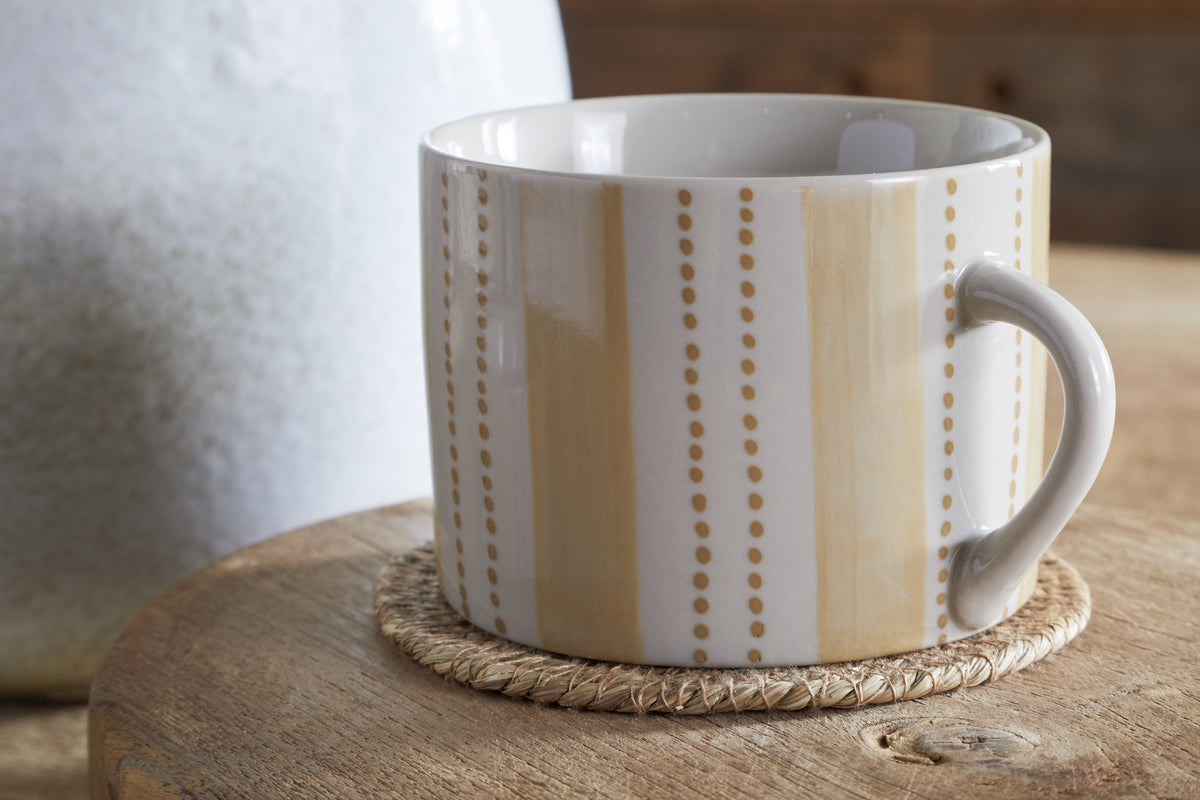 The nkuku Patthar Mug - Cream & Sand (Large, Set of 2) features vertical beige stripes and dotted lines. Hand-painted ceramic mugs rest on a woven coaster atop a round wooden table, with a softly blurred background for rustic charm.