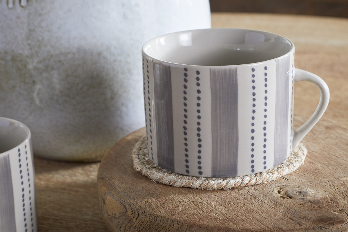 A nkuku Patthar Mug - Cream & Grey - Large (Set of 2) with vertical grey stripes and dotted lines rests on a round woven coaster atop wood, with another hand-painted mug from the collection softly visible in the background.