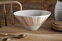 The nkuku Patthar Serving Bowl - Cream & Clay - Medium, hand-painted with beige stripes and dotted lines, rests on a wooden table. A gold spoon with a wrapped handle and a textured plant pot add rustic dinnerware accents.