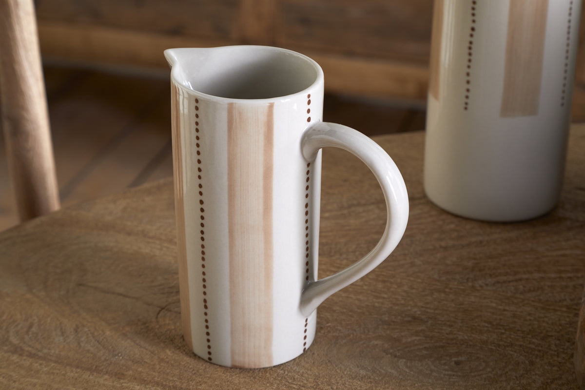 The nkuku Patthar Jug - Cream & Clay - Small, with a tall cylindrical shape, curved handle, and hand-painted tan stripes with dotted lines, sits on a wooden table. A matching jug adds to the warm, rustic background.