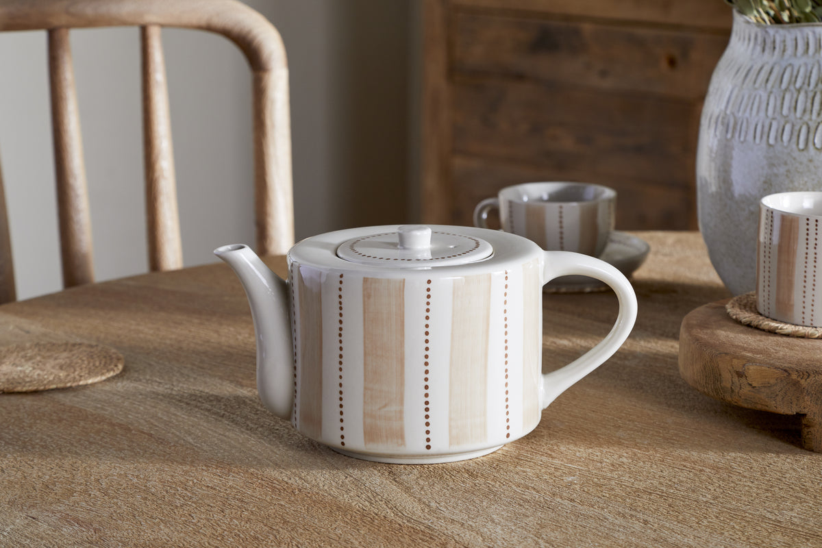 The nkuku Patthar Teapot – Cream & Clay – Large, featuring hand-painted stripes and dots in beige, sits on a wooden table with its matching cup, woven coaster, gray vase with greenery, and wooden chairs in the background.