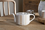 The nkuku Patthar Teapot – Cream & Clay – Large, featuring hand-painted stripes and dots in beige, sits on a wooden table with its matching cup, woven coaster, gray vase with greenery, and wooden chairs in the background.