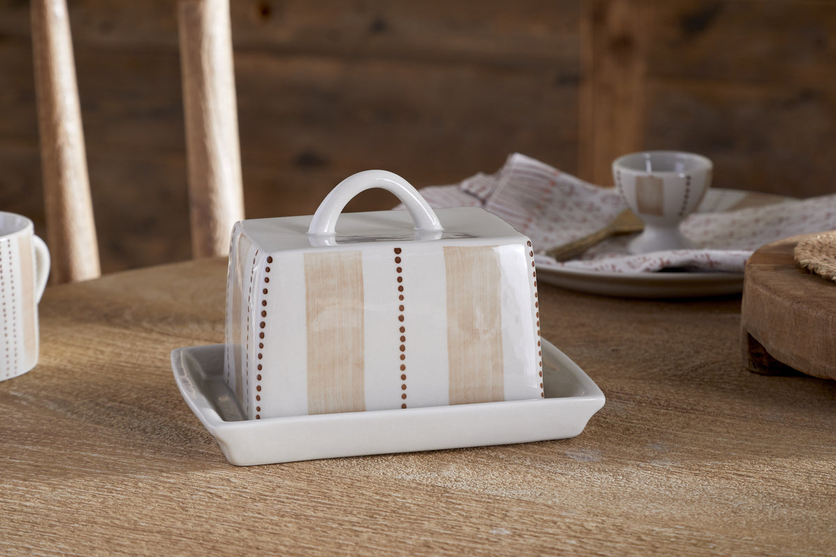 The Patthar Butter Dish - Cream & Clay by nkuku, hand-painted with vertical beige stripes and dotted lines, sits on a wooden table. Dishes, a napkin, and a wooden chair complete the cozy, rustic kitchen scene.