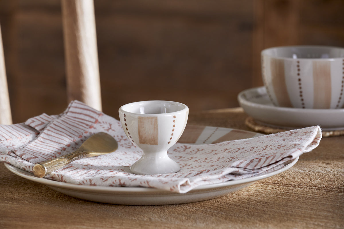 A Patthar Egg Cup from nkuku in cream & clay rests on a plate with a patterned napkin and gold spoon, while its matching bowl adds to the modern, neutral look of this table setting.