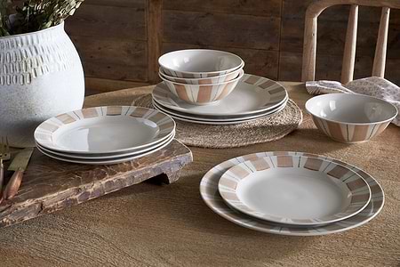 A rustic wooden table is set with nkuku’s Patthar Dinnerware Set - Cream & Clay (set of 12), some dishes stacked, others arranged. A textured white vase with dried flowers and a wooden cutting board add to the cozy dining scene.