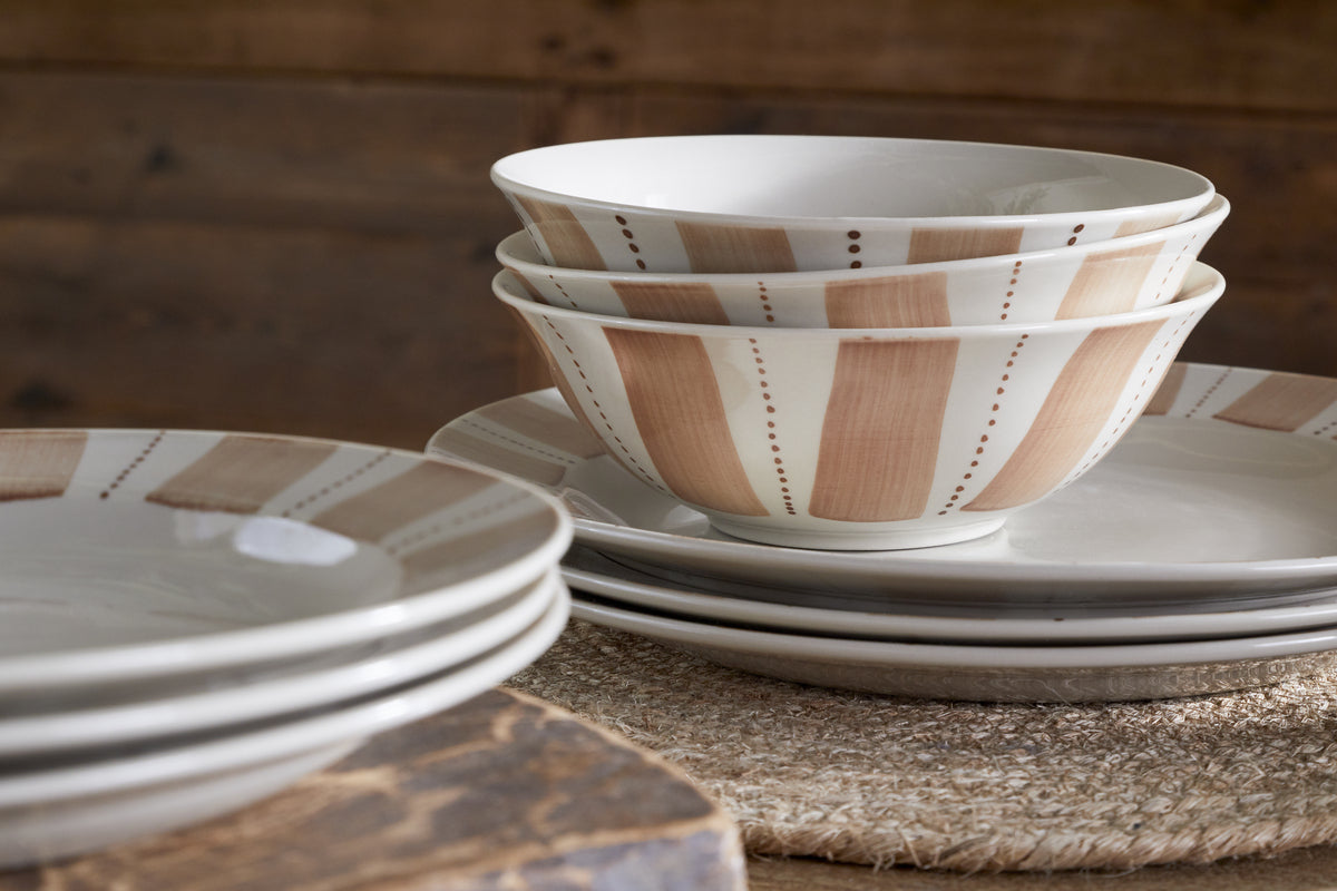A close-up of the nkuku Patthar Dinnerware Set - Cream & Clay (Set of 12) showcases hand-painted beige and brown patterns, stacked on a woven placemat atop wood for a rustic, cozy vibe.