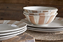 A close-up of the nkuku Patthar Dinnerware Set - Cream & Clay (Set of 12) showcases hand-painted beige and brown patterns, stacked on a woven placemat atop wood for a rustic, cozy vibe.